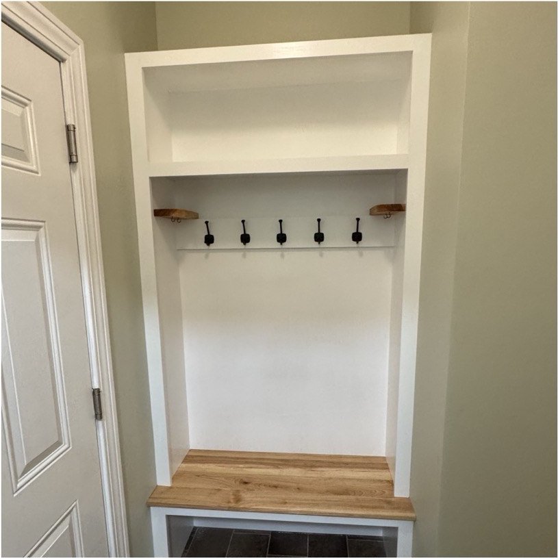 A finished mudroom built-in: a hardwood bench seat in an alcove, a white hook rail with black iron hooks above it, small floating valet trays at each end, and an upper shelf.